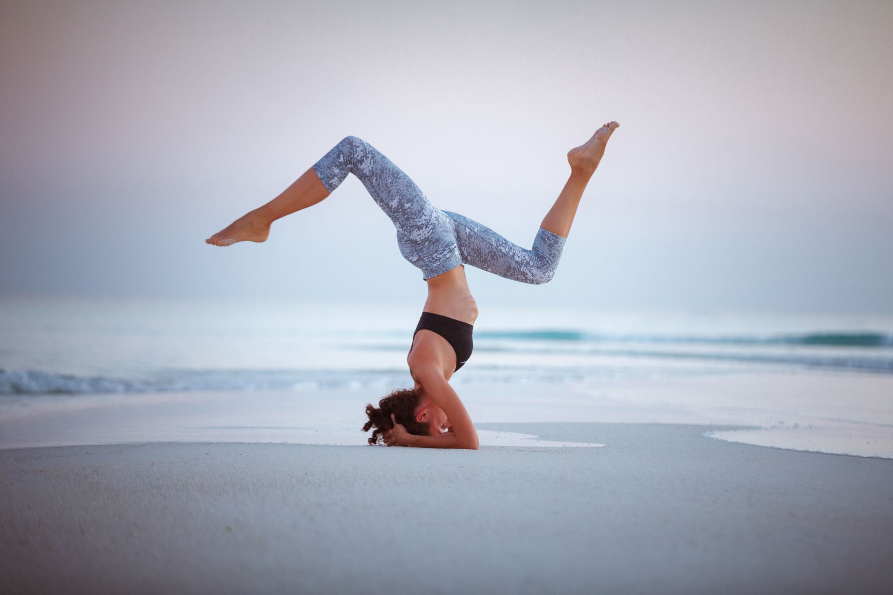A woman performing an advanced headstand yoga pose on a serene, sandy beach at dusk, highlighting the wellness activities and relaxation options available to guests staying with Travel Resort Services.