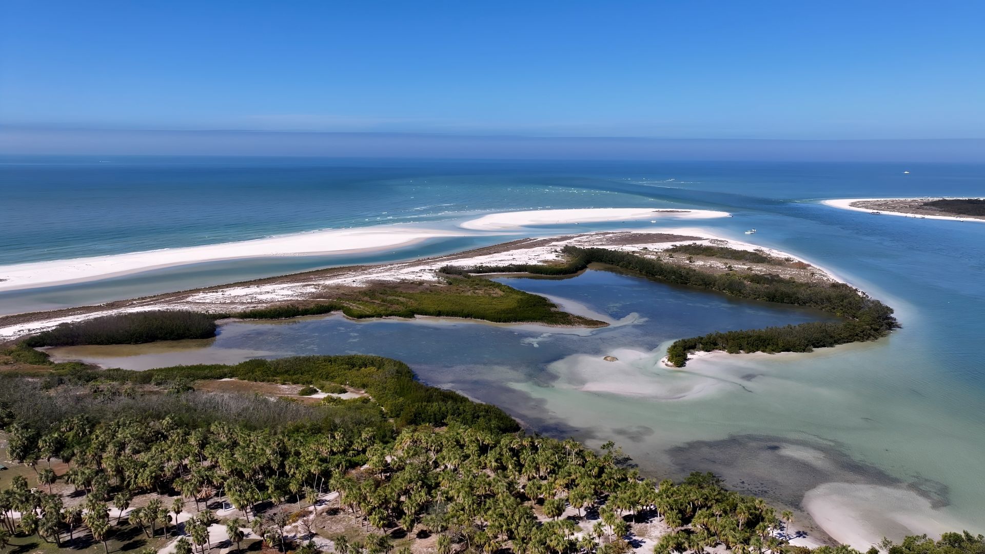 Aerial view of a tropical coastline with white sandbars forming a shallow lagoon and a foreground of palm trees under a blue sky.