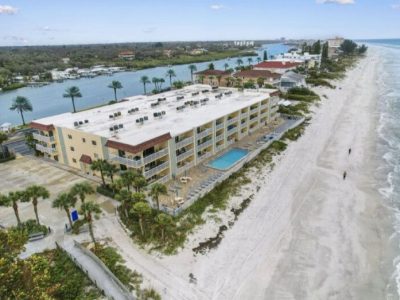 Aerial view of a beachfront hotel with a rectangular pool, palm trees, and a wide white-sand beach along the ocean.