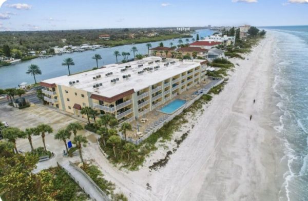 Aerial view of a beachfront hotel with a rectangular pool, palm trees, and a wide white-sand beach along the ocean.