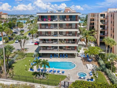 Six-story hotel with balconies, a curved central pool, and lounge chairs amid palm trees under a blue sky.