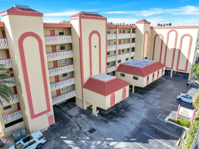 Bird's-eye view of a beige apartment complex with red trim and arched window accents, surrounding a paved parking area under a bright blue sky.