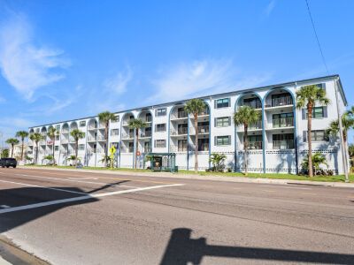 Three-story white apartment building with arched balconies, palm trees lining the sidewalk, and a sunny blue sky.