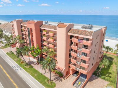 Aerial view of a pink, multi-story seaside apartment complex with palm trees along a road and a sandy beach nearby the ocean