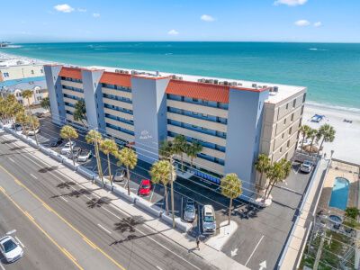 A beachfront hotel building with blue and beige facade, palm trees lining the street, and the ocean in the background.